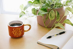 Open notebook and pen next to a mug and plant on a white desk